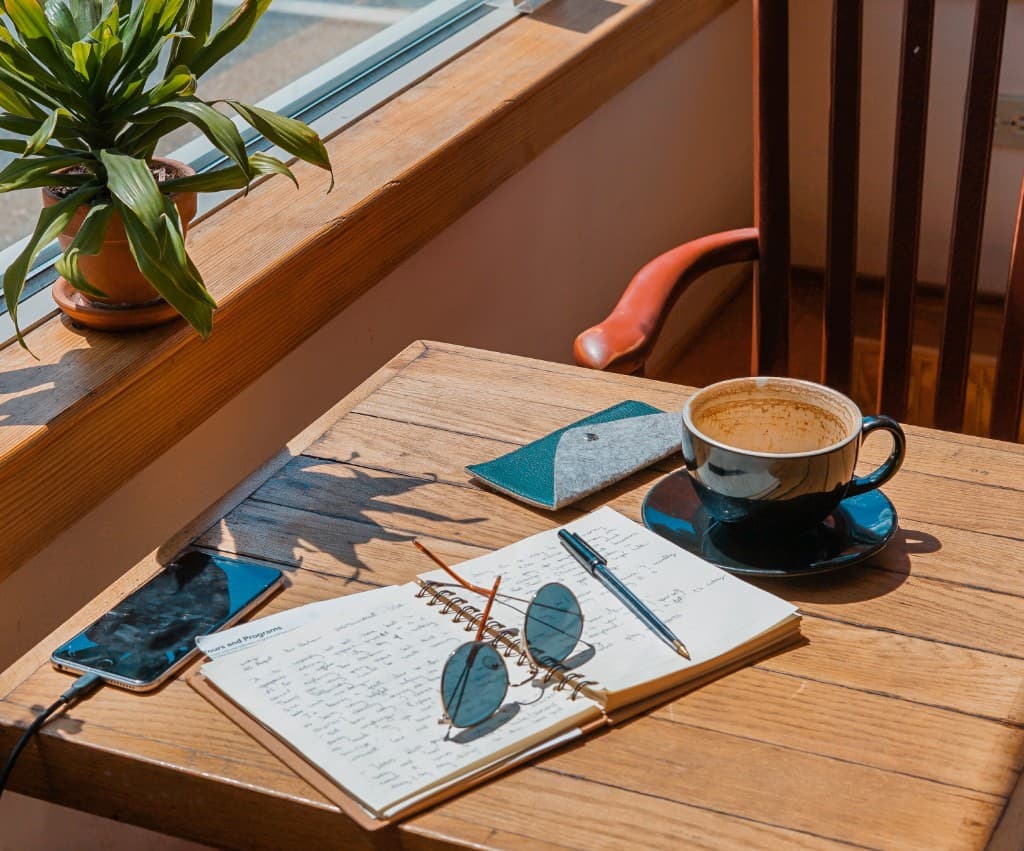 Notebook and coffee on a table by a sunny window.