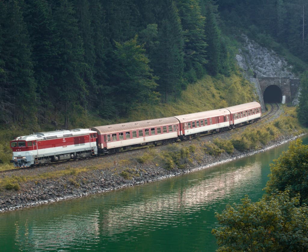 A passenger train traveling beside a forest and river.